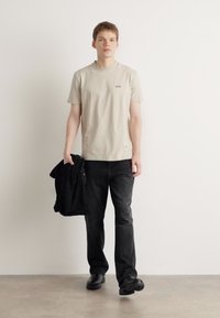 Lyse beige t-shirt med logo, parret med sorte jeans. Personen holder en sort jakke og har sorte sko på. Neutral baggrund.