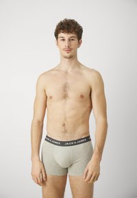 Graue Baumwollboxershorts mit einem schwarzen Bund, der weißen Text trägt. Modell mit lockigem Haar, steht mit entspannten Armen an den Seiten.