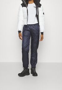 Vit puffjacka med svarta detaljer, tillsammans med marinblå vadderade byxor och svarta stövlar. Klädseln har en åtsittande vit topp under.