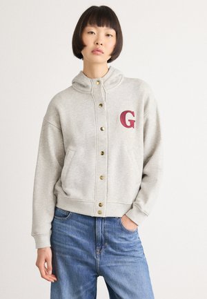 Ungdom med kort mørkt hår iført en lys grå hettegenser med knapper og et rødt "G" på brystet, samt blå jeans og hånden i lommen.