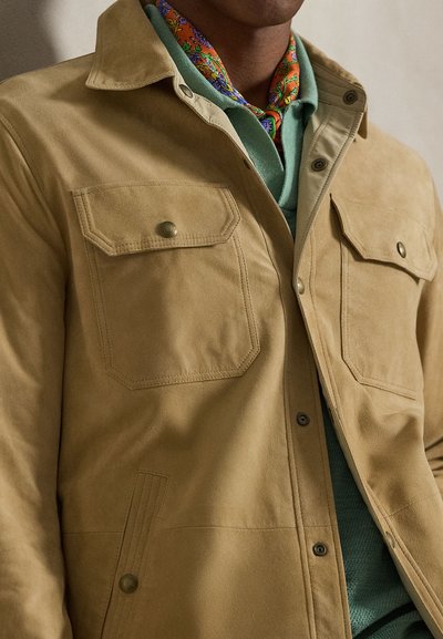 Ταφτά δερμάτινο μπουφάν με δύο τσέπες στο στήθος και κούμπωμα με κουμπιά. Φορεμένο πάνω από ένα πράσινο πουλόβερ, συμπληρωμένο με ένα πολύχρωμο φουλάρι με σχέδια.