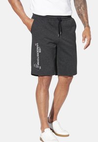 Mann in dunkelgrauen Shorts mit Kordelzug und weißem Text auf dem linken Bein, weißen Turnschuhen und einem weißen T-Shirt vor einem einfarbigen Hintergrund.