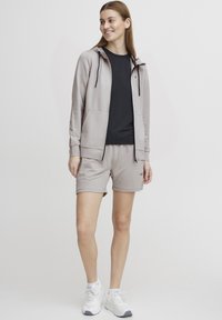 Svijetlosiva hoodie na zip s crnim unutrašnjim dijelom, uparen sa crnom majicom i svijetlosivim šortsevima. Obuća su bijele tenisice.
