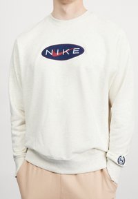 Krémová mikina Nike s kulatým námořnickým logem, na kterém je bílý text a červený swoosh. Ribované manžety a lem, měkká textura.