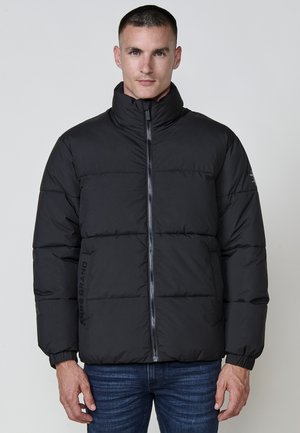 Chaqueta de plumas negra con cuello alto, diseño acolchado y cremallera frontal. Presenta puños elásticos y logotipo en el lado izquierdo.