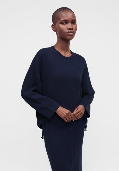 Femme aux cheveux courts portant un pull à manches longues bleu marine foncé et une jupe assortie, debout les mains jointes devant un fond uni.