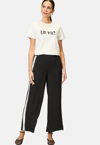 White t-shirt with "ça va?" text in black. Black wide-leg pants with white side stripes. Model wearing black mesh flats, hands at sides.