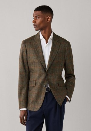 Man staat en kijkt naar links, gekleed in een bruine geruite blazer over een wit overhemd en marineblauwe broek tegen een effen lichte achtergrond.