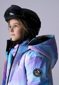 Slēpošanas jaka ar gradientu zilā un violetā krāsā, ar melnu kapuci, melnām detaļām un apaļu logotipa plāksteri uz piedurknes. Valkā melnu ķiveri.