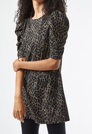 Kobieta ubrana w czarną i złotą sukienkę w abstrakcyjny wzór z bufkami na krótkich rękawach, stojąca z jedną ręką skrzyżowaną na ciele.