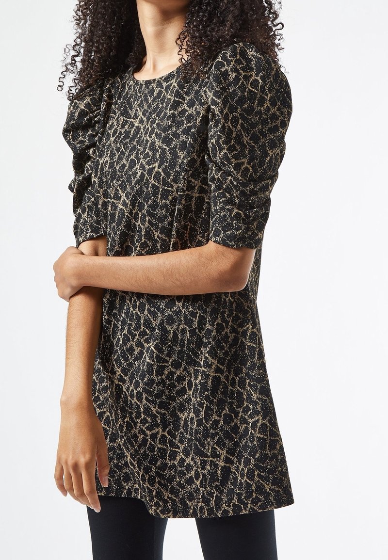 Femme portant une robe noire et or à motif abstrait avec des manches courtes bouffantes, debout avec un bras croisé sur le corps.