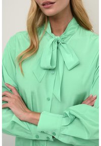Blusa verde menta con un fiocco annodato al collo, chiusura con bottoni e maniche svasate con polsini abbottonati. Tessuto morbido.