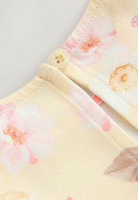 Virágmintás blúzszövet halvány sárga alapon, rózsaszín és narancssárga virágmintákkal. Kis kör alakú gomb található a nyakkivágásnál, puha tapintású.