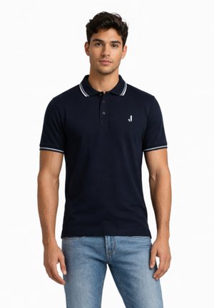 Uomo che indossa una polo blu navy con bordi bianchi su colletto e maniche, abbinata a jeans azzurri, in piedi contro uno sfondo bianco.