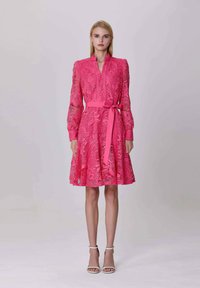 Femme portant une robe en dentelle rose vif avec des manches longues et un nœud à la taille, se tenant contre un fond clair.