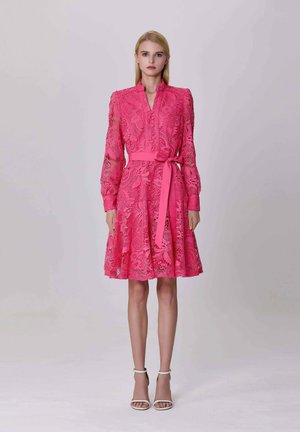 Femme portant une robe en dentelle rose vif avec des manches longues et un nœud à la taille, se tenant contre un fond clair.