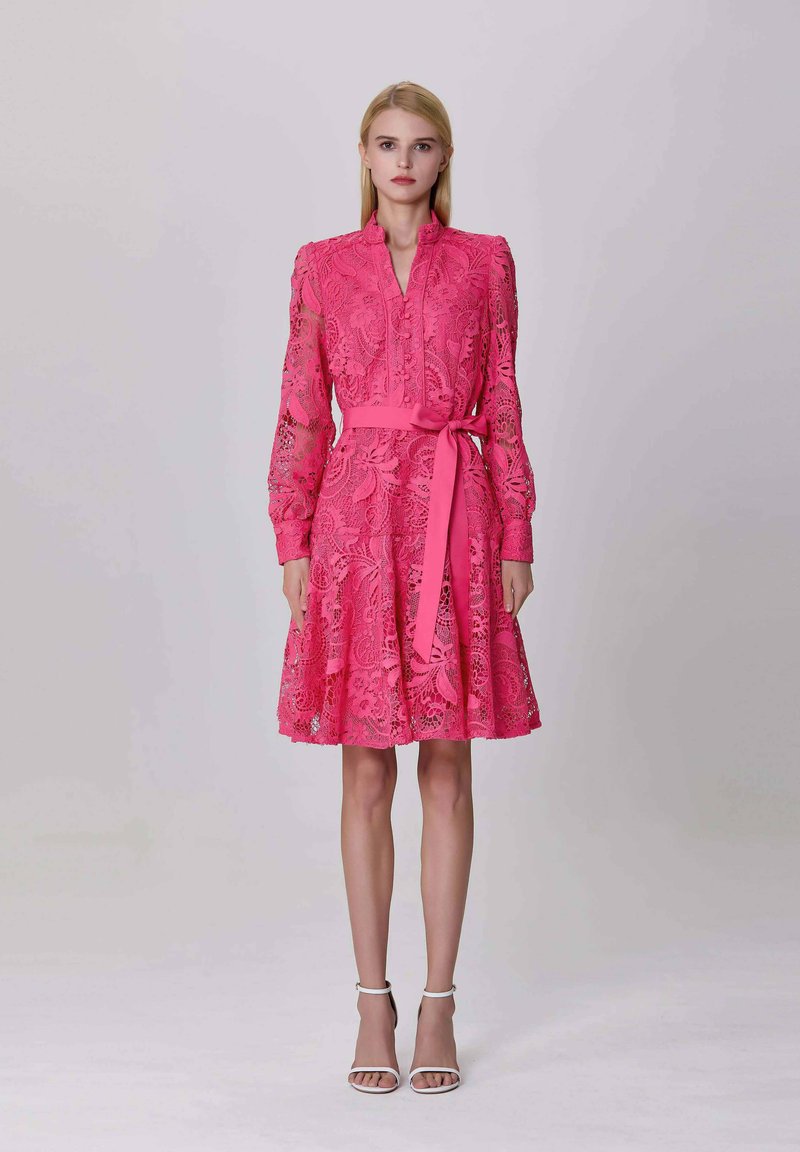 Femme portant une robe en dentelle rose vif avec des manches longues et un nœud à la taille, se tenant contre un fond clair.