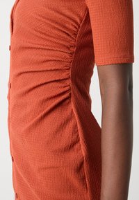Robe couleur rouille avec tissu texturé et détails froncés sur le côté, manches courtes et boutons sur le devant.