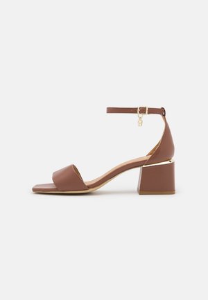 Sandale marron à bout ouvert avec talon bloc, bride cheville avec petite boucle dorée et breloque, et accent doré sur le talon.