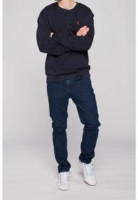 Marinblå sweatshirt med röd logotyp, rundhalsad och ribbade mudd, parat med mörkblå jeans och vita sneakers, som står i en neutral miljö.