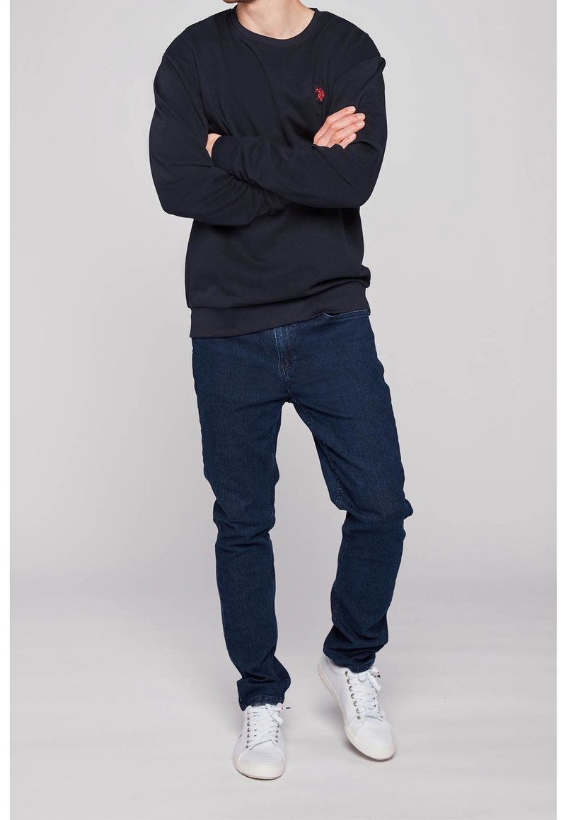 Marinblå sweatshirt med röd logotyp, rundhalsad och ribbade mudd, parat med mörkblå jeans och vita sneakers, som står i en neutral miljö.