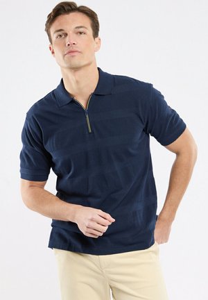 Jeune homme portant un polo bleu marine à manches courtes avec une fermeture éclair au quart et un pantalon beige clair, posant avec une main sur la hanche.