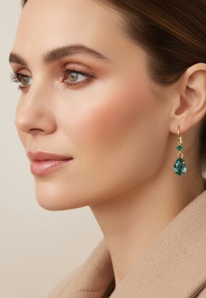 Gouden creolen met een druppelvormige turkoois steen en een kleine groene accentsteen; het model toont ze tegen een neutrale achtergrond.