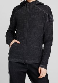 Czarna bluza rozpinana z teksturowanym splotem, kapturem i bocznymi kieszeniami. Posiada subtelne paski i logo Adidasa na górnej lewej stronie.