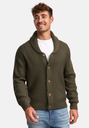 Cardigan de punto verde oscuro con cuello de chal, botones de madera, puños acanalados y tejido texturizado, llevado sobre una camisa blanca.