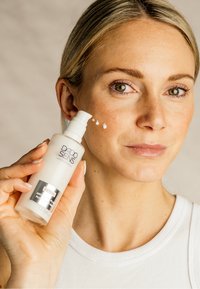 Weiße Flasche der DADO SENS Creme mit Pumpenspender, gehalten neben dem Gesicht einer Frau, die eine glatte, leicht texturierte Creme auf ihrer Haut zeigt.