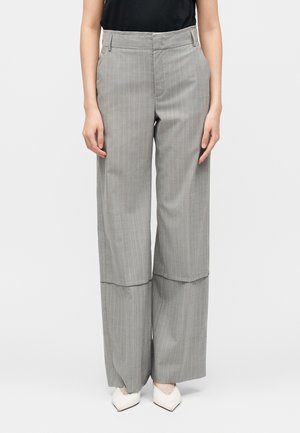 Femme portant un pantalon large gris à fines rayures avec un ourlet superposé sur des talons blancs pointus, associé à un haut noir sans manches.