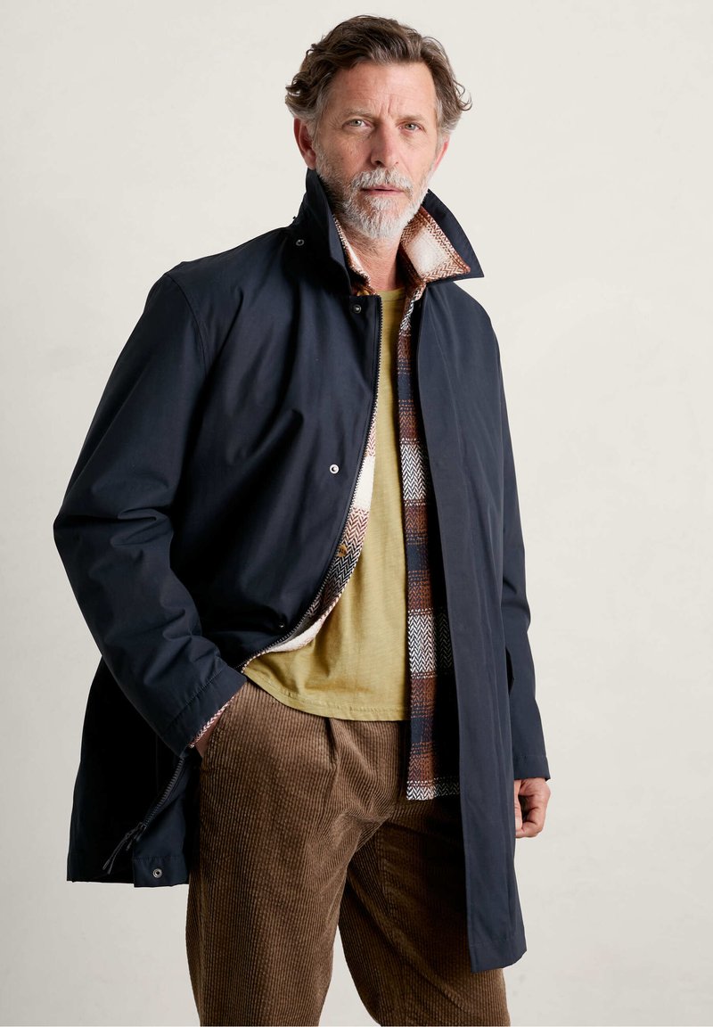 Abrigo impermeable azul marino con un alto cuello, con un forro de cuadros, llevado sobre una camisa mostaza y pantalones de pana marrón.