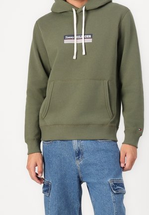 Mężczyzna ubrany w oliwkowo-zieloną bluzę z kapturem Tommy Hilfiger z przednią kieszenią i białymi sznurkami, w połączeniu z niebieskimi jeansowymi spodniami bojówkami.
