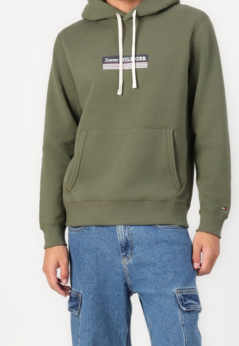 Homme portant un sweat à capuche Tommy Hilfiger vert olive avec poche avant et cordons blancs, associé à un pantalon cargo en denim bleu.