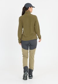 Olivgrön fleecejacka med hög krage, kombinerad med grå och beige tvåtonade byxor. Hållbart material med stövlar. Minimalistisk design utan synliga logotyper.
