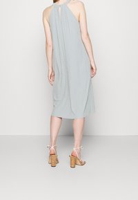 Vestido gris claro sin mangas con un cuello halter torsionado y detalle de abertura en la espalda. La tela tiene una apariencia texturizada y arrugada. Combinado con sandalias de tacón de corcho.