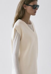 Femme en pull sans manches crème à col en V porté sur un top à manches longues, portant des lunettes de soleil rectangulaires noires, vue de profil sur un fond uni.