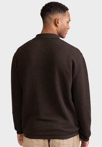 Jersey marrón texturizado con un patrón de ondas, que incluye un cuello y puños acanalados. El diseño es sencillo, con un corte holgado.