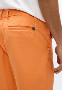 Κοντινή λήψη ενός ατόμου που φοράει πορτοκαλί παντελόνια με πίσω τσέπη που διαθέτει μια μικρή μαύρη ετικέτα λογότυπου Puma στη δεξιά πλευρά.