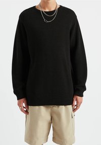 Personne portant un pull noir à côtes, un short beige avec poche latérale, des colliers en argent superposés et des bagues aux deux mains, sur un fond uni.