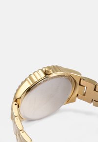 Montre dorée avec un boîtier rond, un cadran texturé et un bracelet en métal. Présente un verre lisse et une couronne pour l'ajustement.