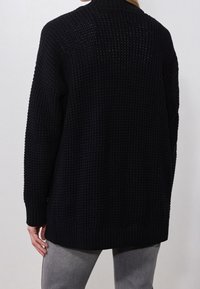 Kvinna som bär en lös svart texturerad stickad tröja och grå byxor, sedd bakifrån mot en enkel ljus bakgrund.