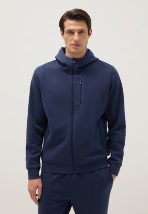 Man draagt een marineblauwe hoodie met rits en bijpassende broek, staat met één hand in de zak tegen een effen lichte achtergrond.