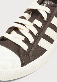 Εγγύς πλάνο καφέ αθλητικού παπουτσιού Adidas με κρεμ κορδόνια, λευκή σόλα, υφή στην μύτη και τρεις κρεμ ρίγες στο πλάι.