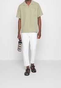 Chemise légère à manches courtes vert clair avec une poche, pantalon blanc resserré, sandales tissées noires et sac à motif noir et beige.
