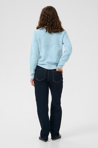 Hellblauer Pullover mit einem strukturierten Muster, kombiniert mit dunklen Jeans, die über Standardtaschen und einen geraden Schnitt verfügen.