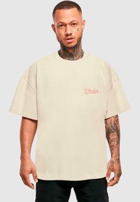 Camiseta beige de gran tamaño con mangas cortas, que presenta un pequeño logo rojo de "2Y Studios" en el pecho y una suave textura de algodón.