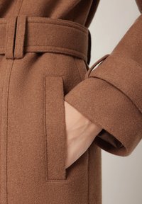 Brązowy wełniany płaszcz z paskiem, charakteryzujący się miękką fakturą, boczną kieszenią i zawiniętym mankietem. Tkanina wydaje się ciepła i dobrze skrojona.