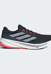 Zapatilla deportiva negra con parte superior de malla, tres franjas plateadas, acentos rojos y una entresuela blanca. Presenta una suela texturizada para mejor tracción.