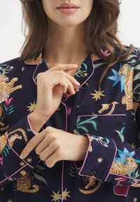 Pigiama blu navy con motivo di tigri e fiori, finiture rosa, dettagli con bottoni e taschino sul petto. Il tessuto appare liscio e leggero.